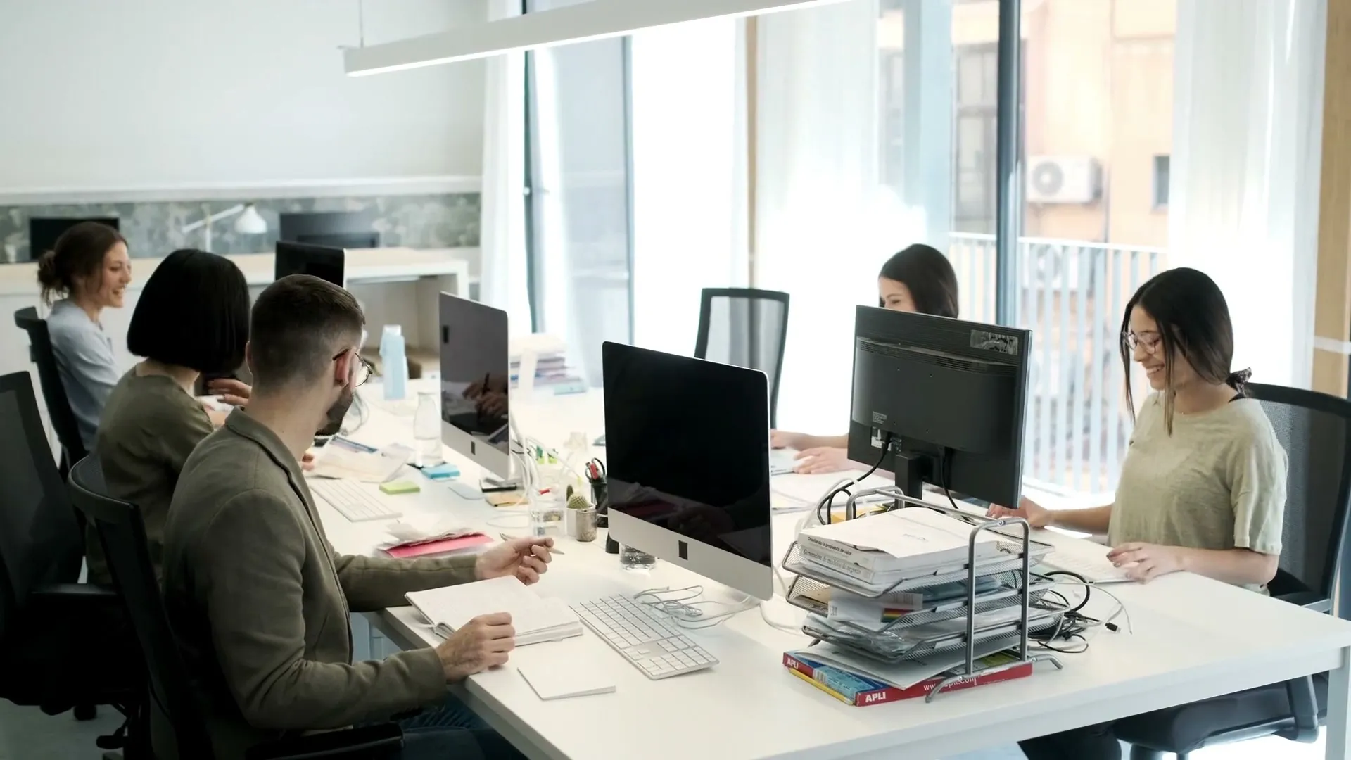 Ein Team an gemeinsamen Schreibtischen in einem hellen Großraumbüro, bei der Arbeit mit Monitoren, Unterlagen und Notizen