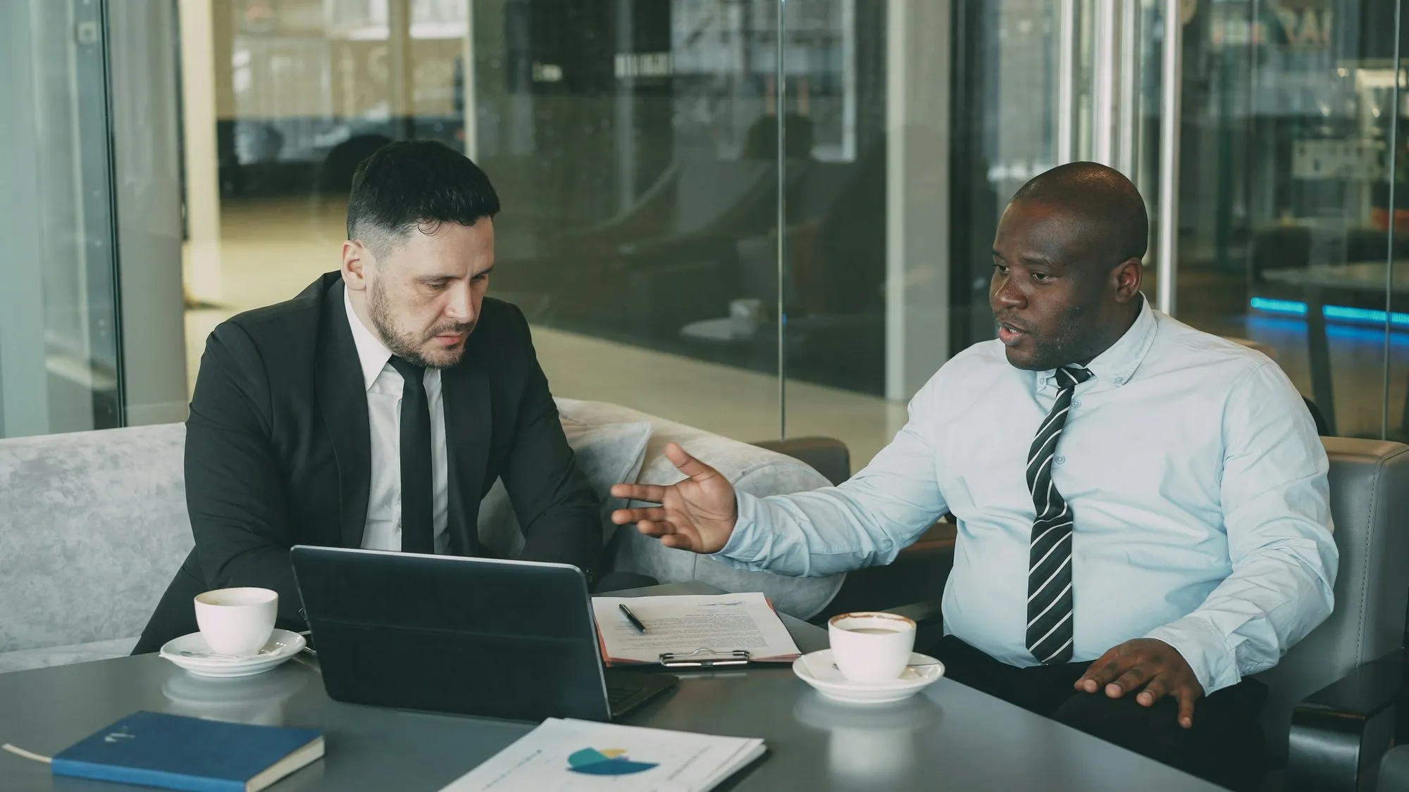 Two businessmen at a table with coffee, a laptop and documents, one of them explaining a point to the other